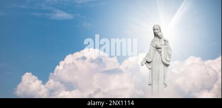 Statue von Mutter Maria und Baby Jesus vor blauem Himmel mit weißen Wolken im Hintergrund. Stockfoto