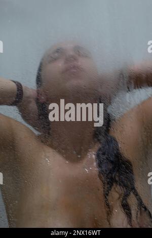 Junge braune, schwarzhaarige Frau unter der Dusche. Blick durch das Glas Stockfoto