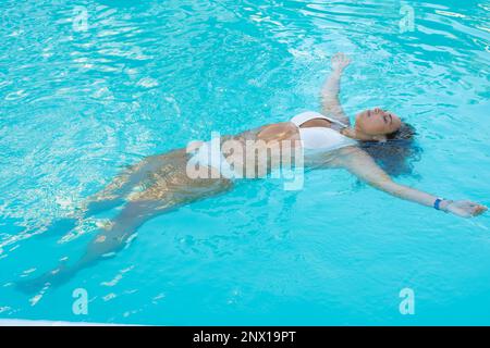 Junge brünette Frau, die einen weißen Bikini in einem Swimmingpool trägt, der im Wasser schwimmt Stockfoto