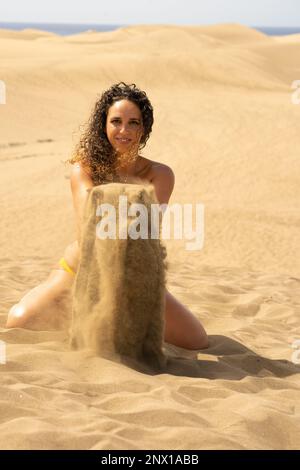 Junge brünette Frau mit lockigem Haar, die mit dem Sand der Dünen von Maspalomas spielt Stockfoto