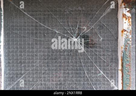 Risse strahlen von einem zentralen Aufschlagpunkt auf einem verstärkten Fenster aus. Stockfoto