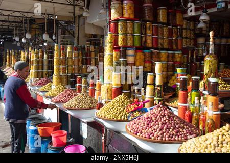 Marrakesch, Marokko – 25. Februar 2023: Ein Mann putzt seine Theke mit Oliven und eingelegtem Gemüse und Obst in Glasgefäßen Stockfoto