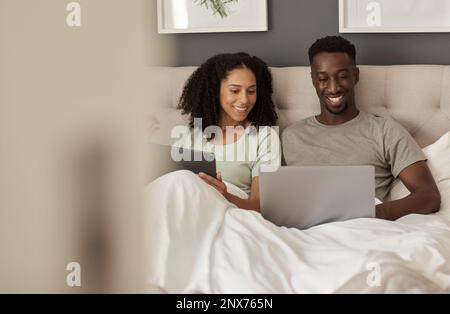 Multiethnisches Paar, das lächelt, während es verschiedene Geräte in seinem Bett verwendet Stockfoto