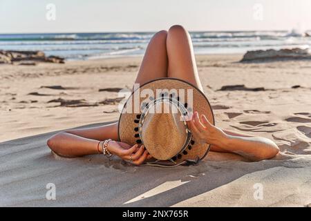 Eine junge Frau entspannt sich an einem Sandstrand und trägt einen braunen Strohhut Stockfoto