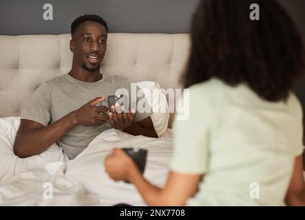 Ein junges, multiethnisches Paar, das beim Kaffee im Bett lächelt und sich unterhält Stockfoto