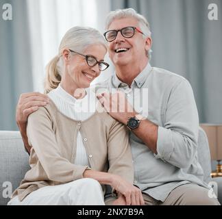 Lachen, lieben oder lustiges altes Paar, das sich auf dem Wohnzimmersofa des Hauses umarmt und eine schöne Zeit genießt. Lächeln, Frieden oder ein glücklicher erwachsener Mann, der sich mit einem verbindet Stockfoto