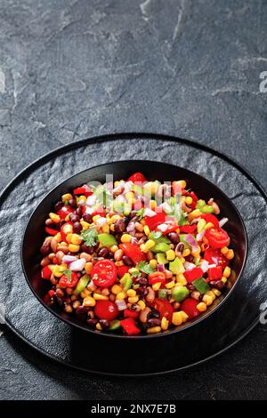 Schwarzer Bohnensalat mit schwarzäugigen Erbsen, Pfeffer, roten Zwiebeln, Mais, Kirschtomaten und Koriander in schwarzer Schüssel, vertikale Ansicht, Nahaufnahme Stockfoto