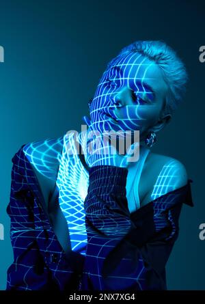 Abstraktes Techno-Foto. Porträt eines jungen blonden Mädchens mit Neonstreifen im Gesicht, das über dunklem Hintergrund in blauen Neonlichtern posiert. Cyberpunk-Stil Stockfoto