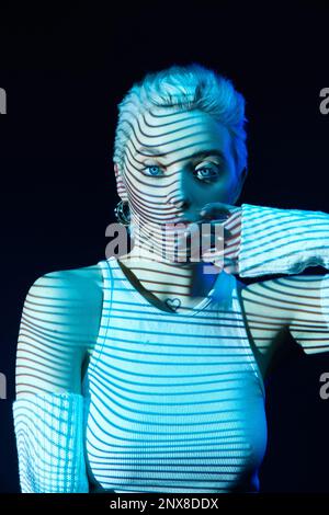 Porträt eines jungen blonden Mädchens mit abstrakten Neonstreifen im Gesicht, das auf dunklem Hintergrund in blauen Neonlichtern posiert. Cyberpunk-Stil Stockfoto