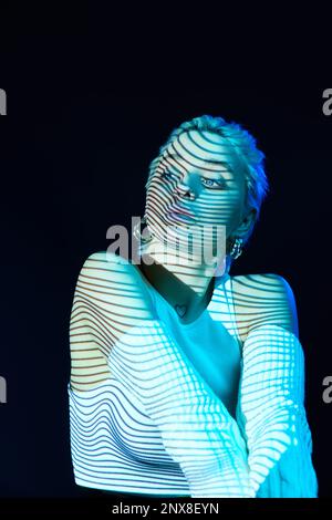 Futuristisch. Porträt eines jungen blonden Mädchens mit Neonstreifen im Gesicht, das über dunklem Hintergrund in blauen Neonlichtern posiert. Cyberpunk-Stil Stockfoto