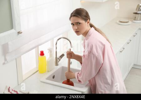 Verärgerte junge Frau, die einen Stößel benutzt, um den Abfluss in der Küche zu reinigen Stockfoto