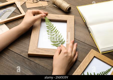 Frau, die wunderschönes Herbarium mit gepresstem, getrocknetem Farnblatt am Holztisch herstellt, Nahaufnahme Stockfoto