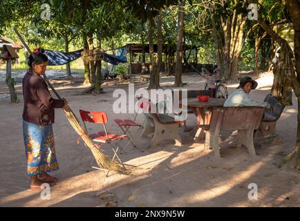 Eine Frau in einem ländlichen Dorf in der Provinz Siem Reap, Kambodscha, fegt Blätter im Innenhof vor ihrem Haus, während sie von einem Nachbarn beobachtet wird. Stockfoto