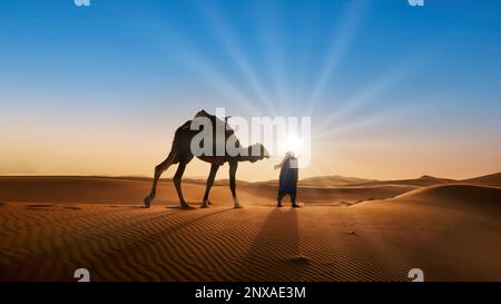 Ein Mann in traditioneller Kleidung wandert bei Sonnenuntergang auf einem Kamel durch eine Wüstenlandschaft. Das Kamel steht auf einer Sanddüne. Der Himmel ist orange, rot. Stockfoto