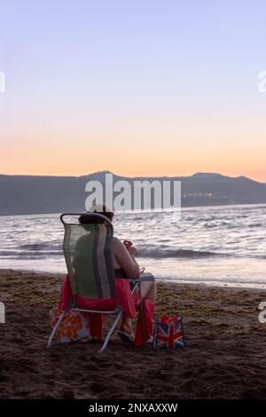 Touristin auf einem Stuhl, die den Sonnenuntergang am Strand mit der Union Jack Flag / Mujer turista en una silla disfrutando de la playa genießt Stockfoto