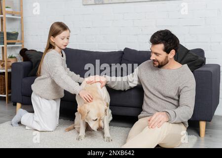 Glücklicher Vater und glückliche Tochter, die auf dem Boden neben der Couch sitzen und labrador streicheln, Bild der Börse Stockfoto