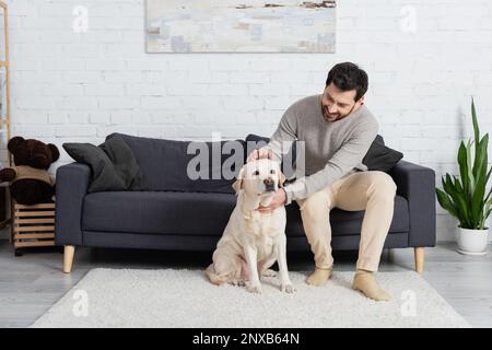 Voller Mann mit fröhlichem Bart, der auf der Couch im Wohnzimmer sitzt und labrador streichelt, Stockbild Stockfoto