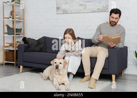 Lächelnder bärtiger Mann liest Buch, während er auf der Couch neben der Tochter saß und labrador-Hund streichelte, Stockbild Stockfoto