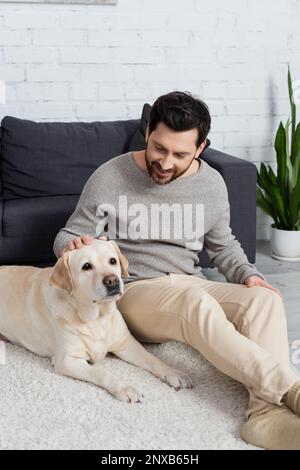 Fröhlicher Mann mit Bart, der auf dem Boden neben dem Sofa sitzt und labrador streichelt, Stockbild Stockfoto