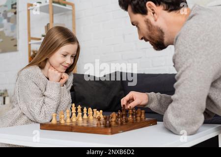 Ein bärtiger Mann bewegt die Schachfigur, während er zu Hause mit einer lächelnden Tochter spielt, Stockbild Stockfoto