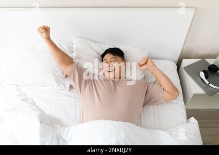 Ein glücklicher asiatischer Mann, der einen guten Morgen genießt, sich im Bett ausstreckt Stockfoto