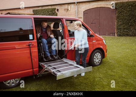 Seniorin hält die Fernbedienung und hilft einem Mann mit Behinderung im motorisierten Rollstuhl beim Aussteigen aus dem Van Stockfoto