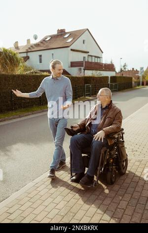 Die ganze Länge eines lächelnden Seniorenmannes mit Behinderung im motorisierten Rollstuhl durch einen jungen Pflegenden, der während der Sonne auf der Straße unterwegs ist Stockfoto