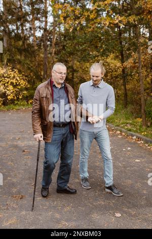 Die ganze Länge eines jungen männlichen Betreuers, der einen pensionierten älteren Mann beim Laufen mit Stock auf der Straße unterstützt Stockfoto