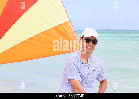 Porträt eines glücklichen Mannes mit Baseballkappe und Sonnenbrille unter Segel auf Seehintergrund. Segelsport, Strandurlaub Stockfoto