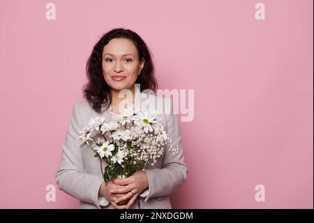 Wunderschöne Frau mittleren Alters, die vor die Kamera lächelt, mit einem Strauß Frühlingsblumen posiert, auf pinkfarbenem Hintergrund Stockfoto