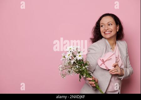 Eine reizende Frau lächelt zur Seite und posiert mit einem Strauß weißer Frühlingsblumen und einer Geschenkbox auf pinkfarbenem Hintergrund Stockfoto