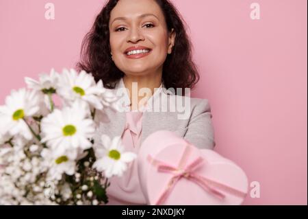 Eine lächelnde Frau hält vor der Kamera einen Strauß weißer Kamillenbäume und eine herzförmige Geschenkbox für den Mutter- oder Frauentag Stockfoto