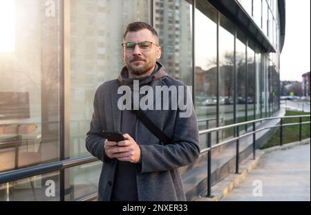 Ein Brillenunternehmer, der neben einem Bürogebäude steht, sein Handy in der Hand hält und lächelt. Stockfoto