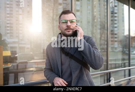 Ein Mann mit Brille und Mantel, der auf der Straße steht und mit dem Handy telefoniert. Stockfoto