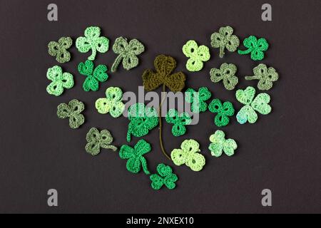Komposition aus gestrickten grünen Shamrocks in Herzform auf schwarzem Hintergrund. Weihnachtsschild und gestricktes Kleeblatt. Kopierbereich, flach liegend, Stockfoto