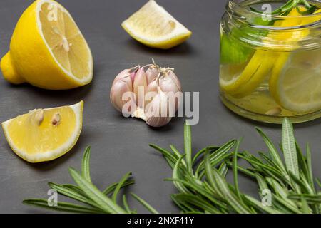 Rosmarinzweige, Knoblauch und Zitrone auf dem Tisch. Zitronengetränk in einem Glasgefäß. Schließen. Grauer Hintergrund. Stockfoto