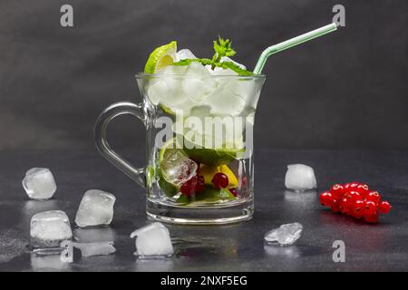 Limette, rote Johannisbeere, Minze, Eis und Strohhalme in einem Glas. Geschmolzenes Eis und ein Zweig rote Johannisbeere auf dem Tisch. Schwarzer Hintergrund. Stockfoto