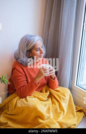 Eine Frau mit weißen Haaren, die am Fenster sitzt und einen heißen Tee zum Entspannen nach einem Arbeitstag trinkt. Konzept: Entspannung, Ruhe, mit mir selbst. Stockfoto