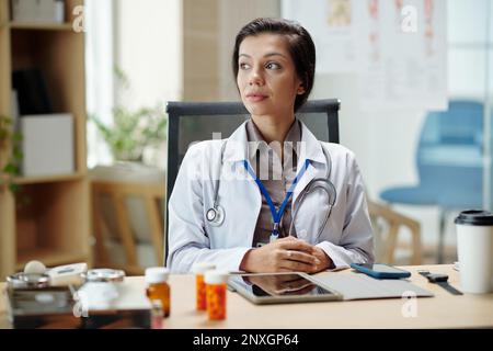 Pensive Leiterin der Medizinischen Klinik sitzt am Bürotisch und schaut weg Stockfoto