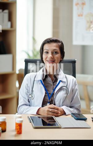 Lächelnde, junge Hausärztin, die an ihrem Schreibtisch sitzt Stockfoto