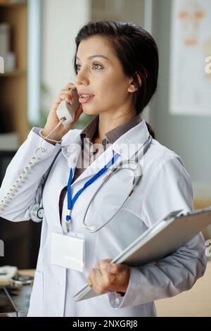 Junge Frau, die in der medizinischen Klinik arbeitet, spricht mit dem Patienten am Telefon Stockfoto