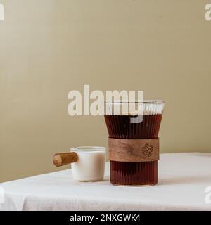 Kaffee in einem durchsichtigen Glas auf dem Tisch, Milch in einem Milchkännchen, Schokolade auf einem Teller. Stockfoto