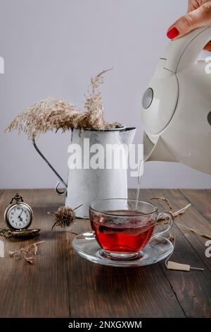 Eine weibliche Hand gießt heißes Wasser aus einem Wasserkocher in einen klaren Glasbecher, um Tee zu machen, mit einer alten Uhr und einer Vase mit trockenen Pflanzen daneben Stockfoto