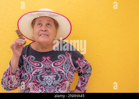 Alte Frau mit einer Zigarre in der Hand vor gelbem Hintergrund, überraschter Gesichtsausdruck, Großmutter trägt einen Hut und genießt den sonnigen Tag. Konzept von Stockfoto