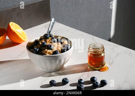 Gesunder Haferbrei mit Blaubeeren oben in weißer Porzellanschüssel, Honigdose und Orangen im Hintergrund Stockfoto