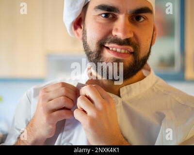 Porträt eines jungen, bärtigen Kochs, der lächelt, während er seine Kochjacke anzieht und in die Kamera schaut Stockfoto