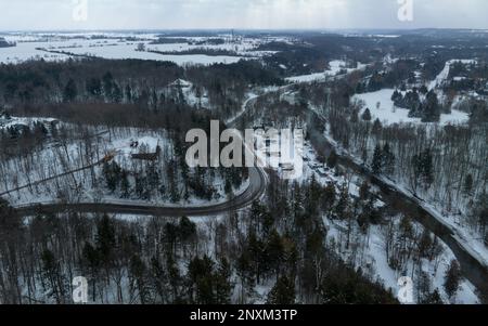 Eine zweispurige Landstraße, die sich durch ein schneebedecktes hügeliges, bewaldetes Gebiet schlängelt, ist an einem verschneiten Wintertag aus der Vogelperspektive zu sehen. Stockfoto