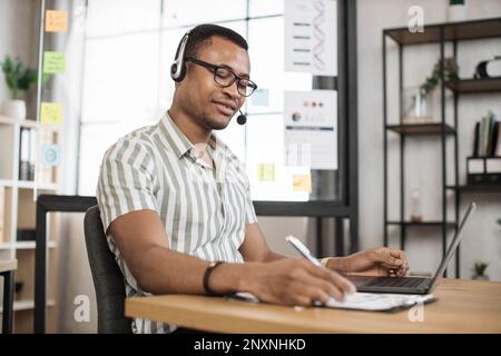 Porträt eines lächelnden jungen afrikanischen Büroleiters in gestreiftem Hemd und Headset, der während der Online-Arbeit mit einem drahtlosen Laptop einen Finanzbericht schreibt. Stockfoto
