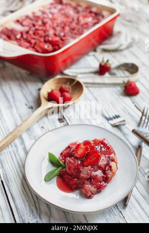 Aus einem hohen Blickwinkel können Sie süßen, hausgemachten Erdbeerkuchen oder Sonker genießen, die in einer roten Keramikpfanne über einem rustikalen weißen Holztisch gebacken wurden. Stockfoto