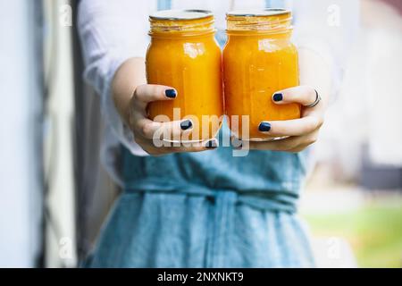 Eine Frau in einer Schürze, die frische hausgemachte Pfirsichbutter hält. Hausbau. Selektiver Fokus auf Maurerglas mit unscharfem Hintergrund. Stockfoto